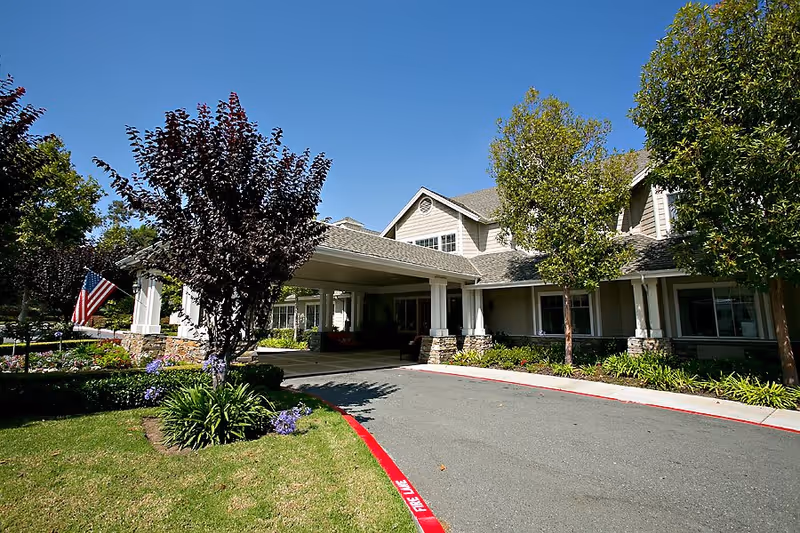 Exterior view of a senior living facility building with a covered entrance driveway, surrounded by trees and landscaping under a clear blue sky.