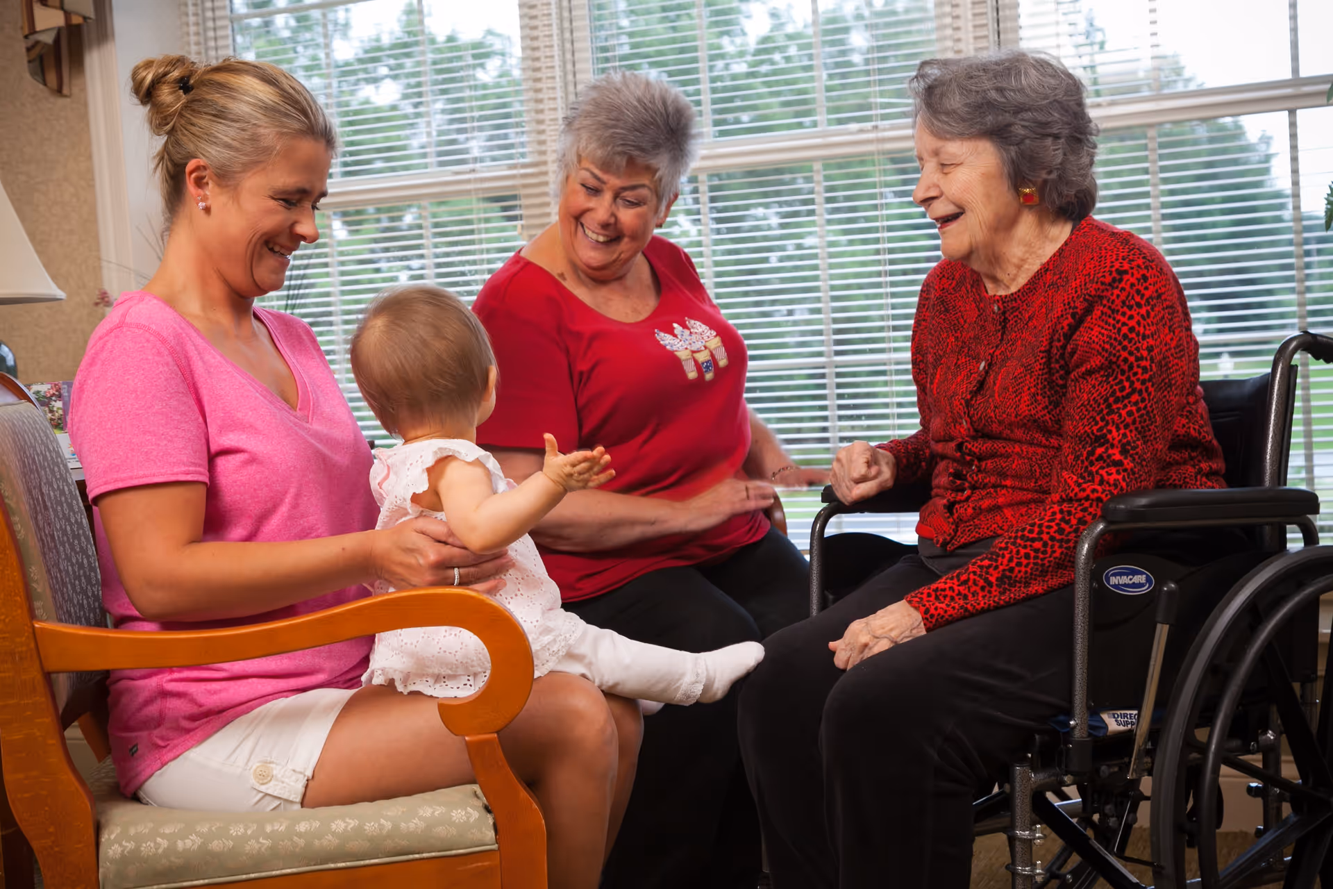 Three generations of women smiling and interacting in a bright room with large windows. A young woman in a pink shirt holds a baby dressed in white, while an elderly woman in a red sweater sits in a wheelchair, and a middle-aged woman in a red shirt looks on happily.