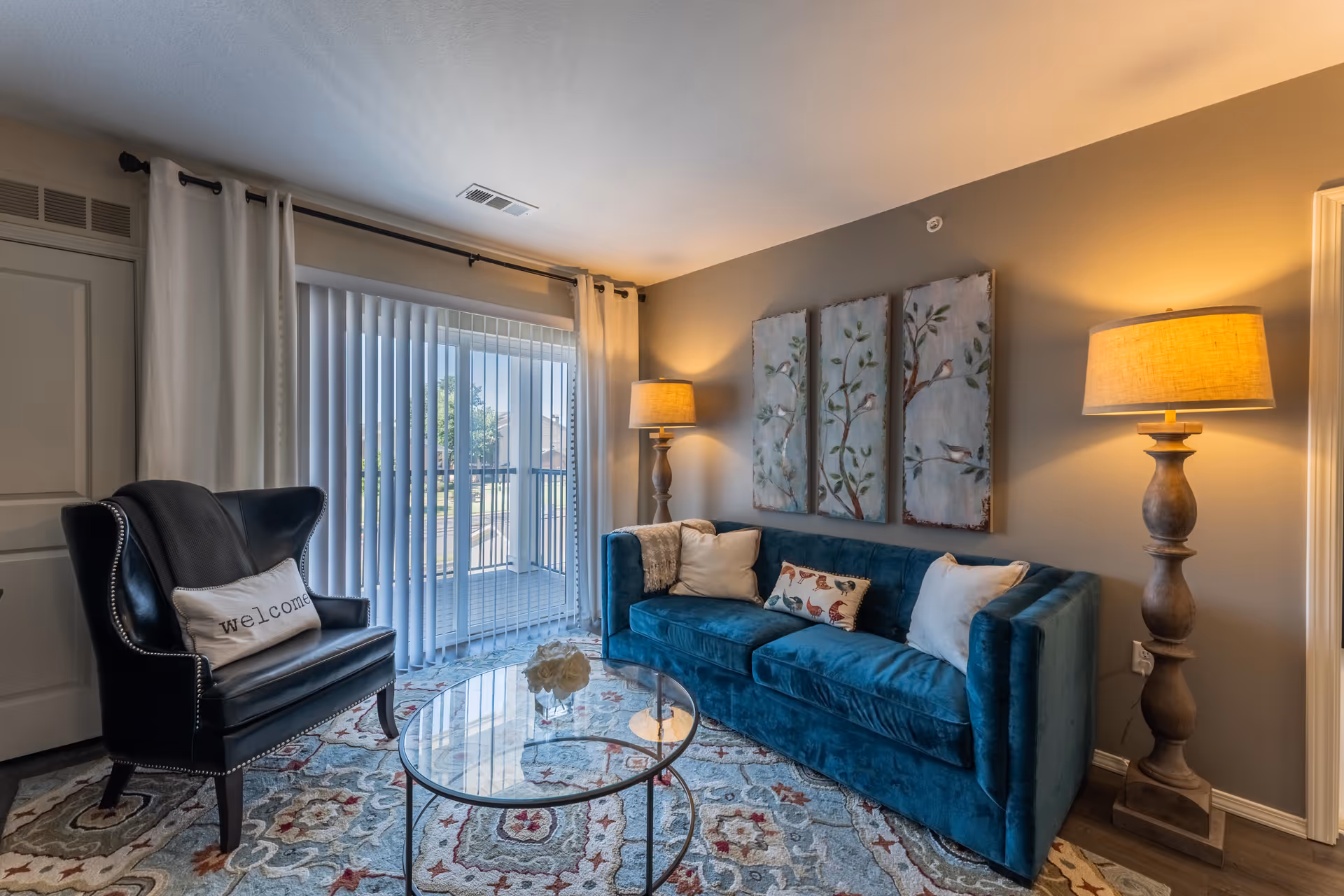 A cozy living room with a blue velvet sofa adorned with decorative pillows, a black armchair with a 'welcome' pillow, a round glass coffee table, two tall wooden floor lamps with beige lampshades, and three nature-themed paintings on the wall. The room has a large window with vertical blinds and white curtains, allowing natural light to enter.