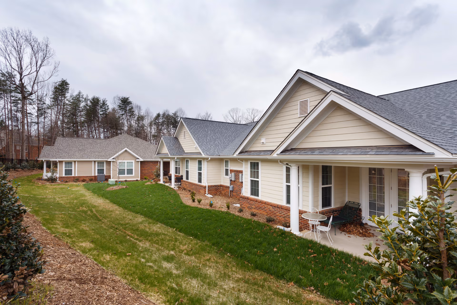 Exterior view of a senior living facility with beige siding and brick accents, featuring a covered porch with outdoor seating. The building is surrounded by a well-maintained lawn and landscaping, with trees in the background under a cloudy sky.