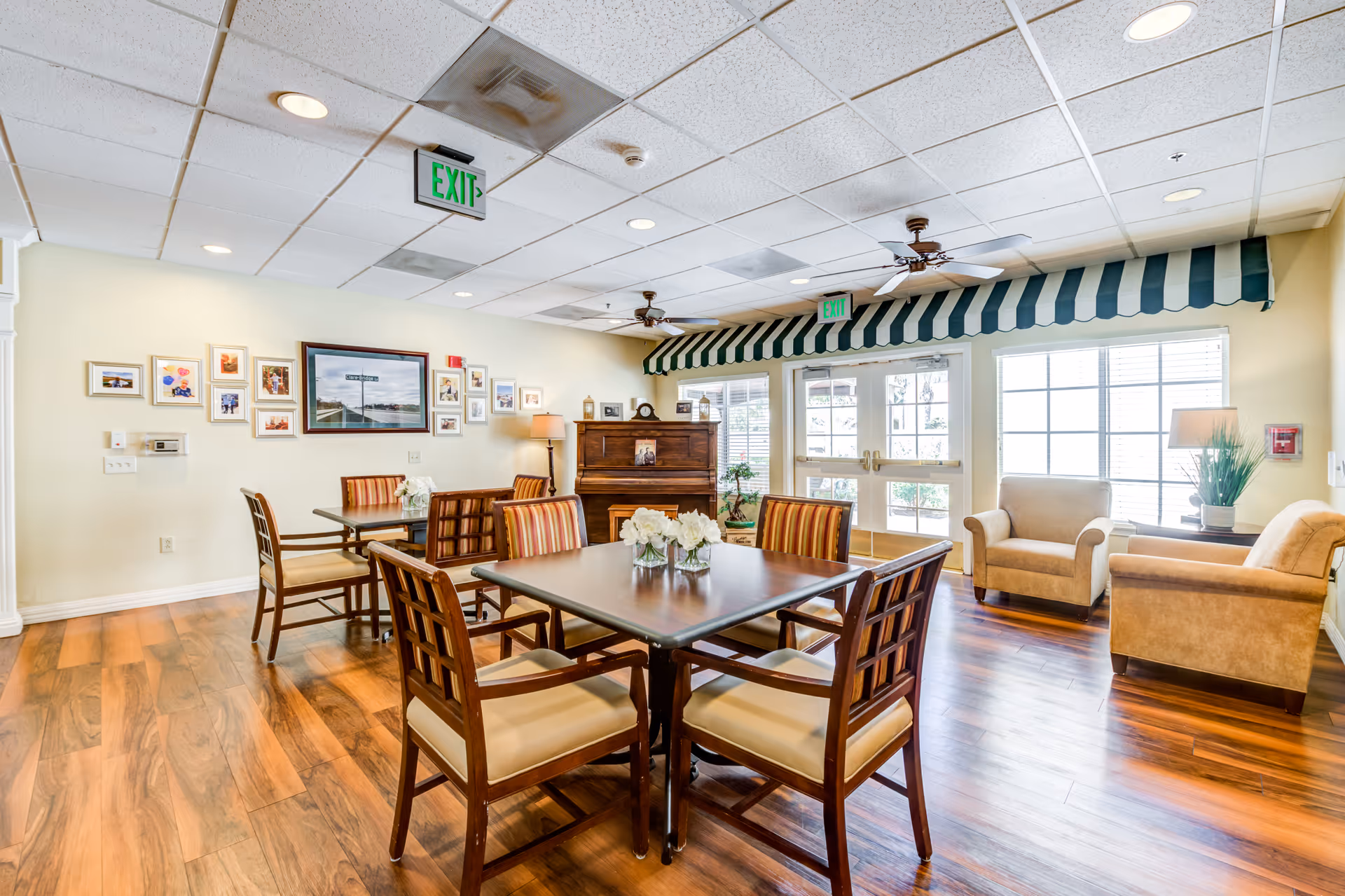 A bright and inviting common area in a senior living facility featuring wooden tables with cushioned chairs arranged for socializing or dining. The room has wood flooring, a piano against the far wall, framed pictures on the wall, two armchairs near large windows, ceiling fans, and exit signs above the doors. A striped green and white awning is above the double doors leading outside.