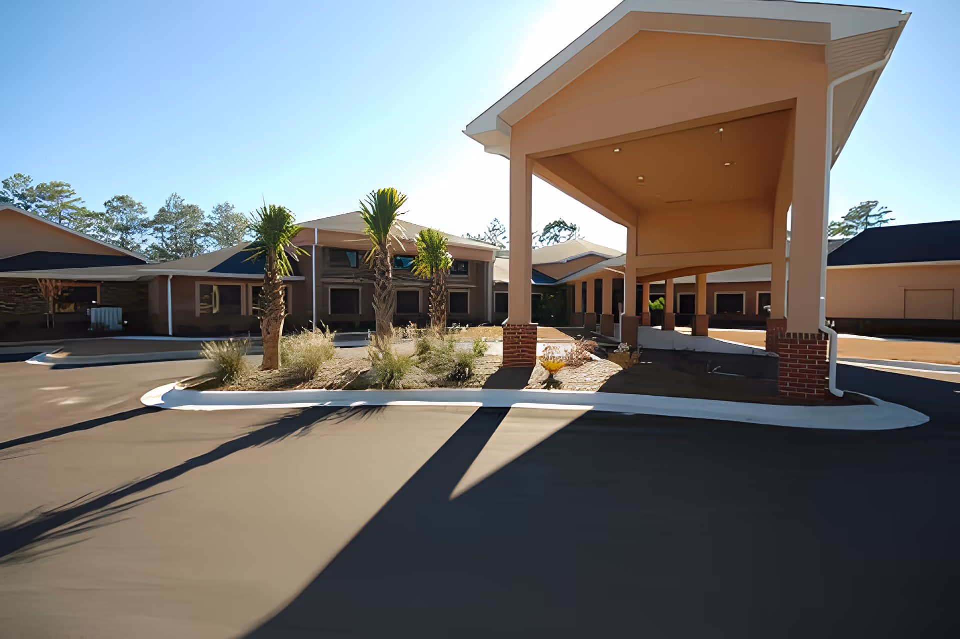Covered driveway and porte-cochere at the front of a single-story senior living building with palm trees and landscaped islands.