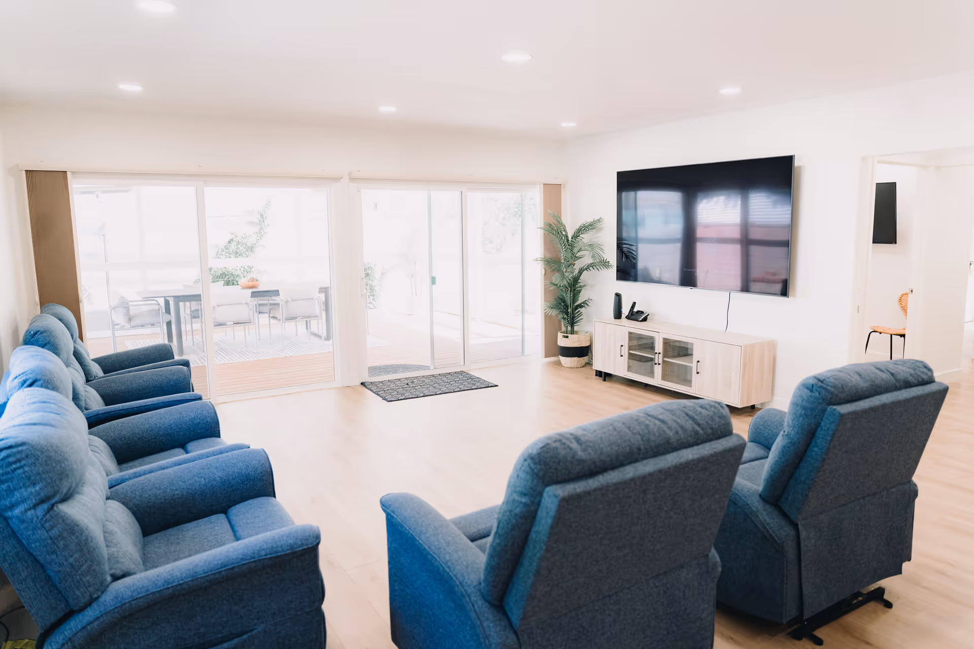 A bright and spacious living room with several blue upholstered recliner chairs arranged facing a large flat-screen TV mounted on the wall. There is a light wood TV stand below the TV and a potted plant next to it. Large sliding glass doors lead to an outdoor patio area with a table and chairs.