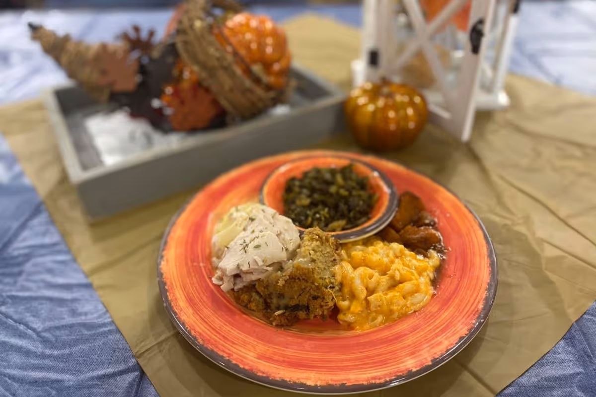 A close-up view of a plate with a serving of macaroni and cheese, cooked greens in a small bowl, roasted sweet potatoes, stuffing, and sliced turkey. The plate is on a table with a brown tablecloth, and there are autumn-themed decorations including small pumpkins and a tray with pine cones in the background.