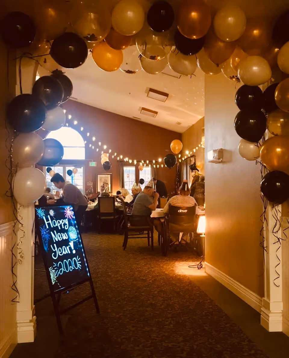 Decorated dining room with balloons, string lights and a chalkboard sign reading 'Happy New Year!' where residents sit at tables for a celebration.