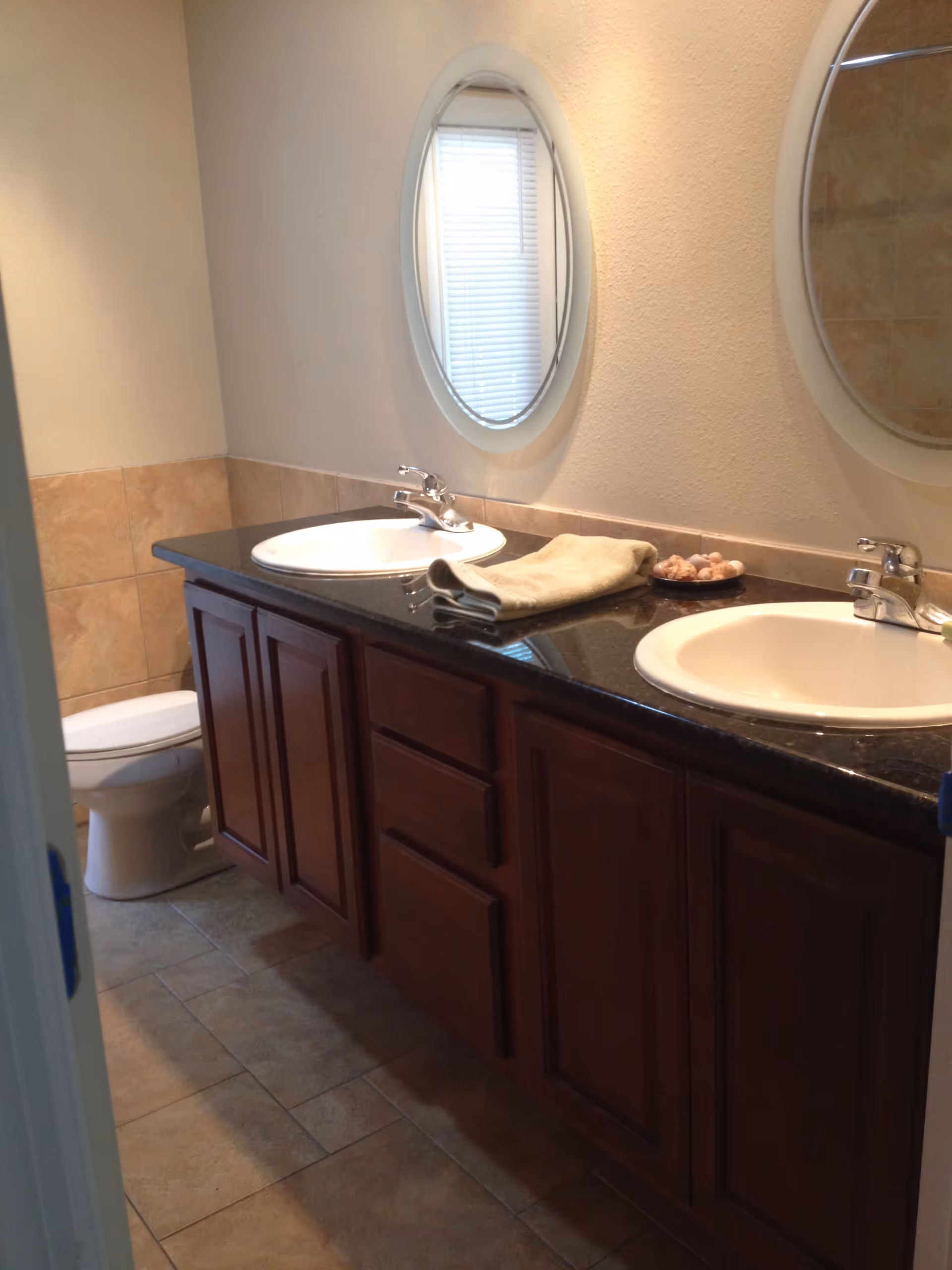 Bathroom with a double sink vanity featuring dark granite countertop and wooden cabinets. Two oval mirrors are mounted on the wall above the sinks. A folded towel and a small dish with decorative items are placed on the countertop. A toilet is visible in the corner next to the vanity. The floor and lower wall are tiled in a beige color.