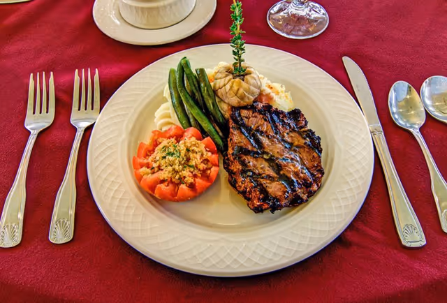 A plated grilled steak with green beans, mashed potatoes topped with roasted garlic, and a stuffed tomato on a white plate set on a red tablecloth with silverware and a glass.