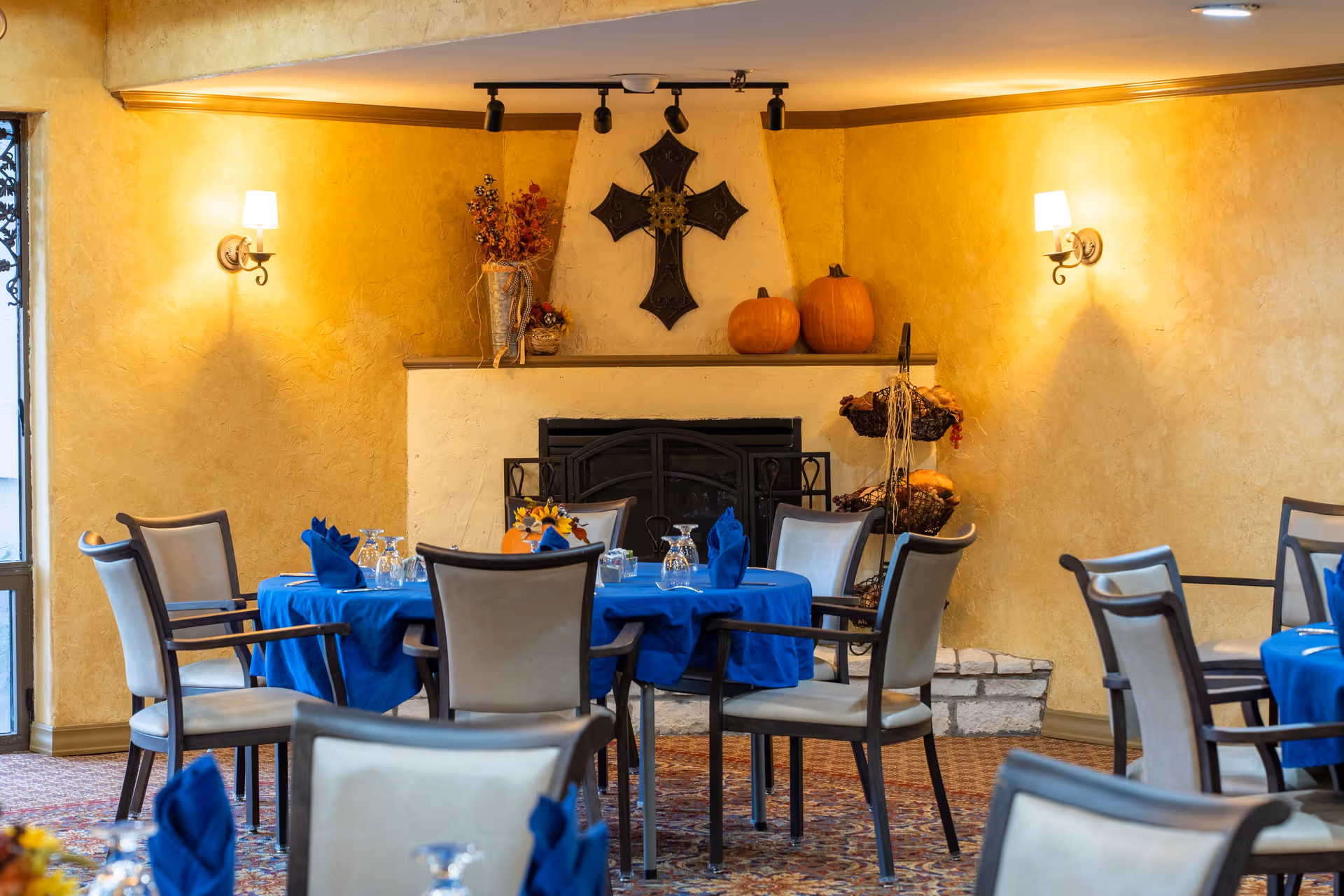 Dining area with round tables covered in blue tablecloths and set with glasses and napkins. The room features a fireplace decorated with pumpkins, a metal cross, and autumn-themed floral arrangements. Warm yellow walls and wall-mounted lamps create a cozy atmosphere.