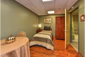 A small bedroom with a single bed covered in a striped comforter and a pillow. There is a wooden wardrobe against the wall, a framed picture above the bed, and a round table with a decorative item on it. The room has wooden flooring and green walls. An open door leads to a bathroom with a shower visible.