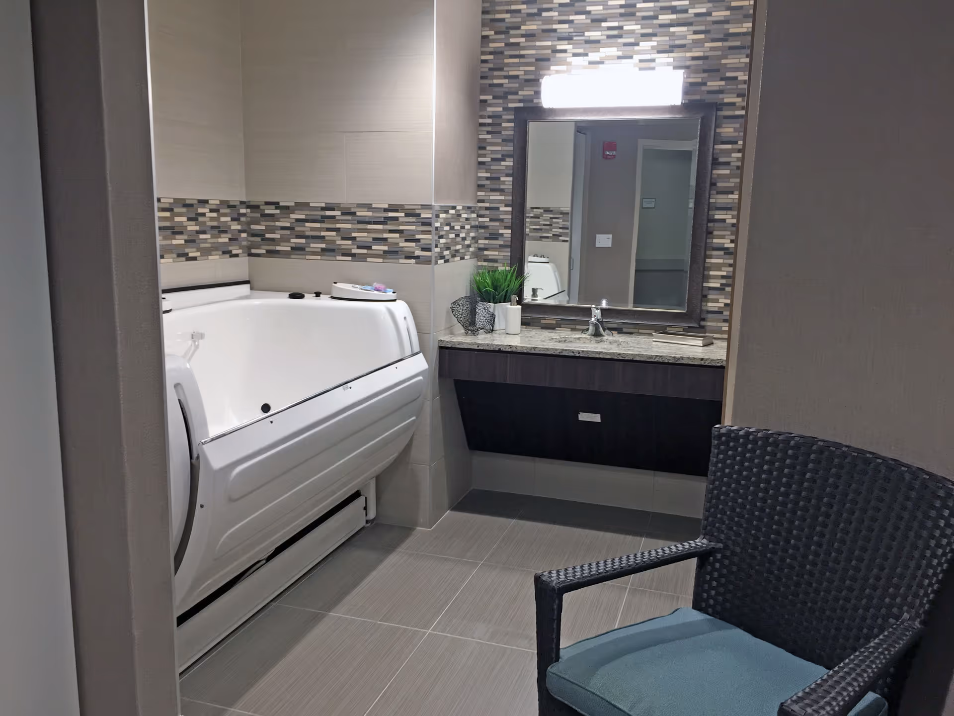 Modern bathroom with a walk-in bathtub, vanity and mirror set against a tiled accent wall, and a wicker chair.