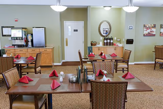 A bright dining room with wooden tables and chairs, each table set with red folded napkins and a beverage station along the back wall.