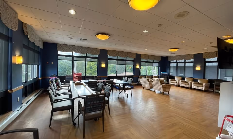A spacious senior living common area with large windows letting in natural light. The room features multiple tables and chairs arranged for group activities, including a shuffleboard table and a Connect Four game. There are also several comfortable armchairs and sofas along the far wall. The walls are painted blue with decorative valances above the windows, and the ceiling has recessed lighting and round ceiling fixtures.