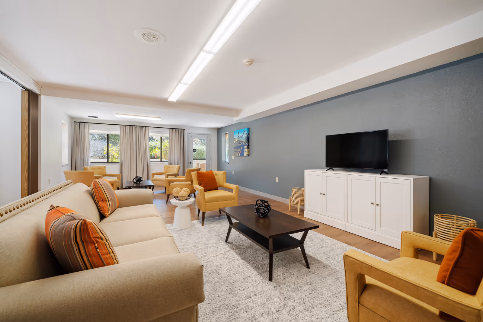 A bright and spacious living room in a retirement community featuring a beige sofa with striped and orange cushions, two yellow armchairs with orange pillows, a dark wooden coffee table on a light patterned rug, a white TV cabinet with a flat-screen television, and large windows with beige curtains allowing natural light to fill the room.