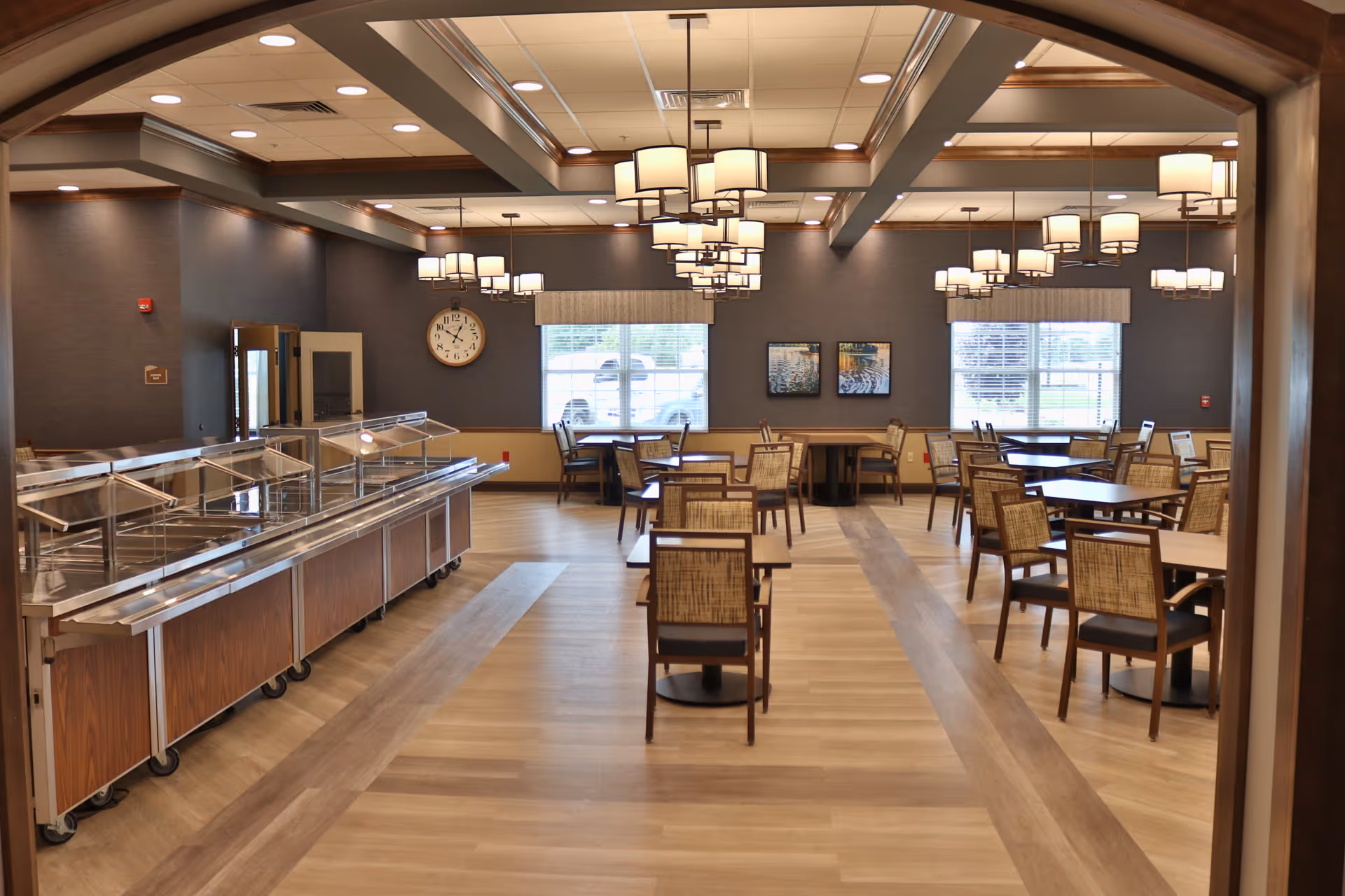 A spacious dining room with multiple wooden tables and chairs arranged neatly. There are large windows with blinds allowing natural light to enter. The ceiling features wooden beams and multiple modern chandeliers providing warm lighting. On the left side, there is a stainless steel buffet serving station with glass sneeze guards. The walls are decorated with a clock and framed artwork.