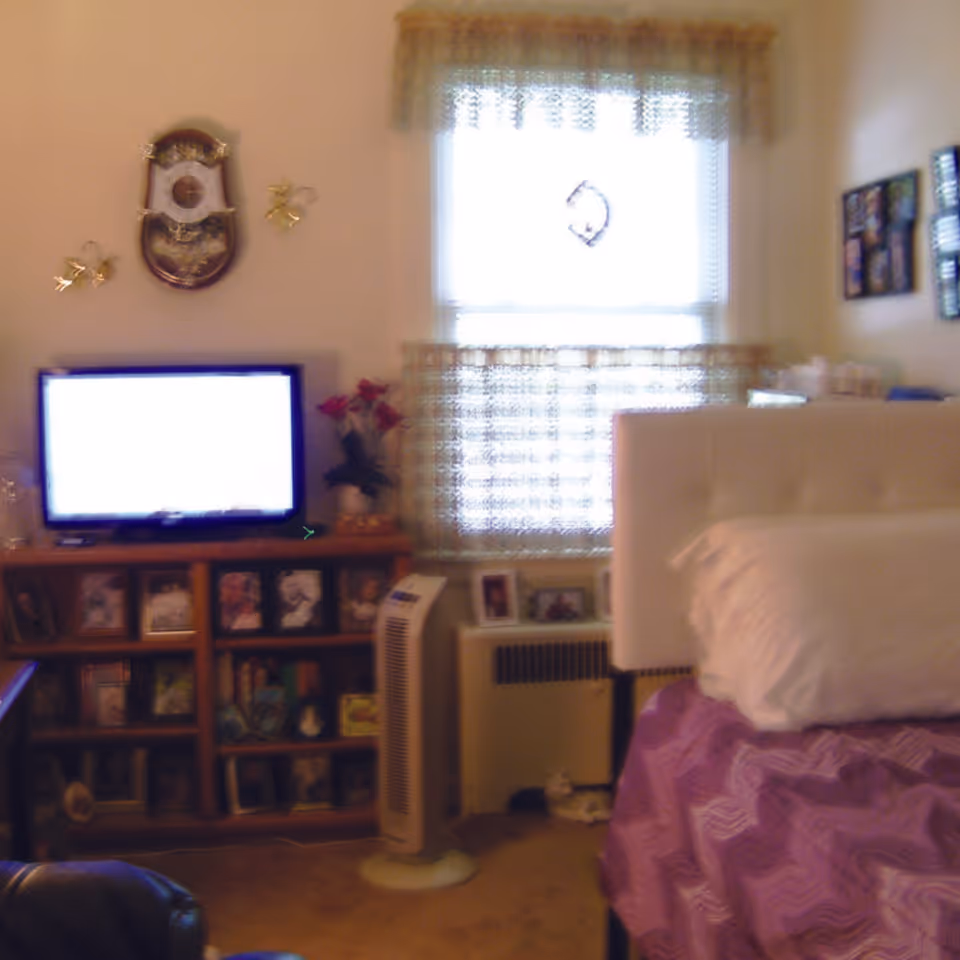 A cozy bedroom with a white tufted headboard and purple bedspread. There is a window with lace curtains letting in light. A wooden shelf unit holds a TV and several framed photos. A tall white fan stands on the floor next to the shelf. The walls have decorative items including a clock and framed pictures.