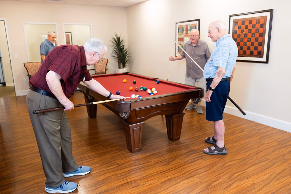 Three elderly men playing pool on a red felt pool table in a well-lit room with wooden flooring. Two men are holding pool cues and one is bent over aiming a shot. In the background, there is a man standing near a doorway and two framed artworks on the wall.