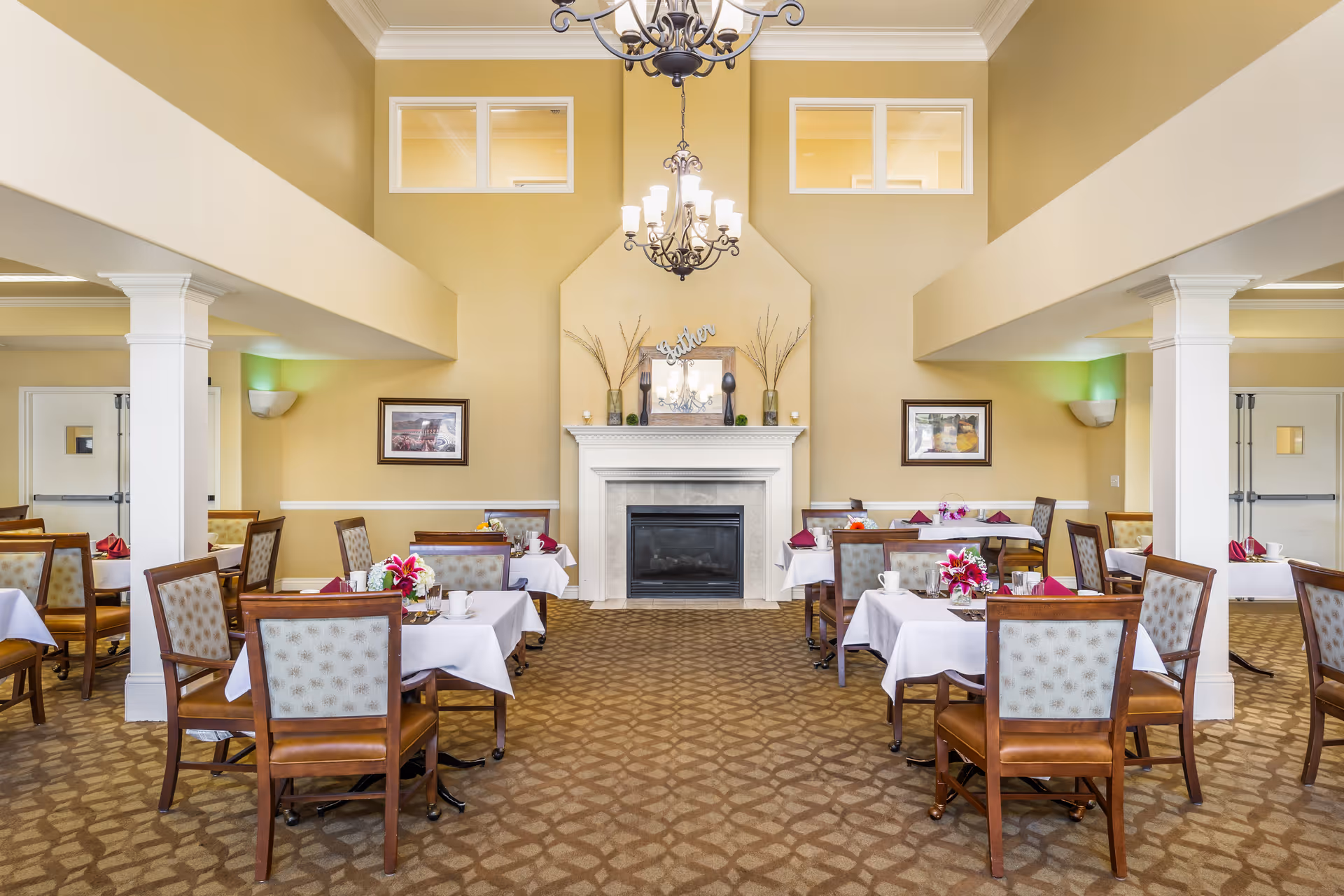 Bright dining room with tables set for meals, upholstered chairs, a central fireplace, and a chandelier.
