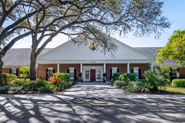Front exterior view of a single-story brick building with white trim and a peaked roof, surrounded by landscaped greenery and large trees casting shadows on the driveway.