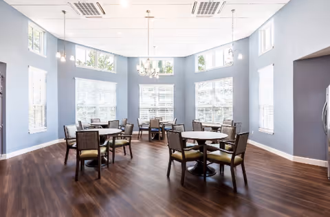 Bright dining room with several round tables and chairs, large windows, and a central chandelier.