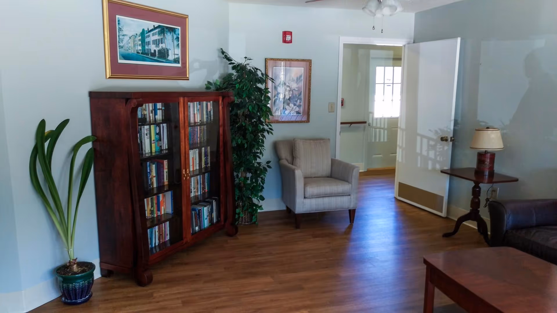 A cozy interior room with wooden flooring featuring a glass-front wooden bookshelf filled with books, a potted plant beside it, a beige armchair, a side table with a lamp, and a partially open door letting in natural light. Two framed pictures hang on the walls.