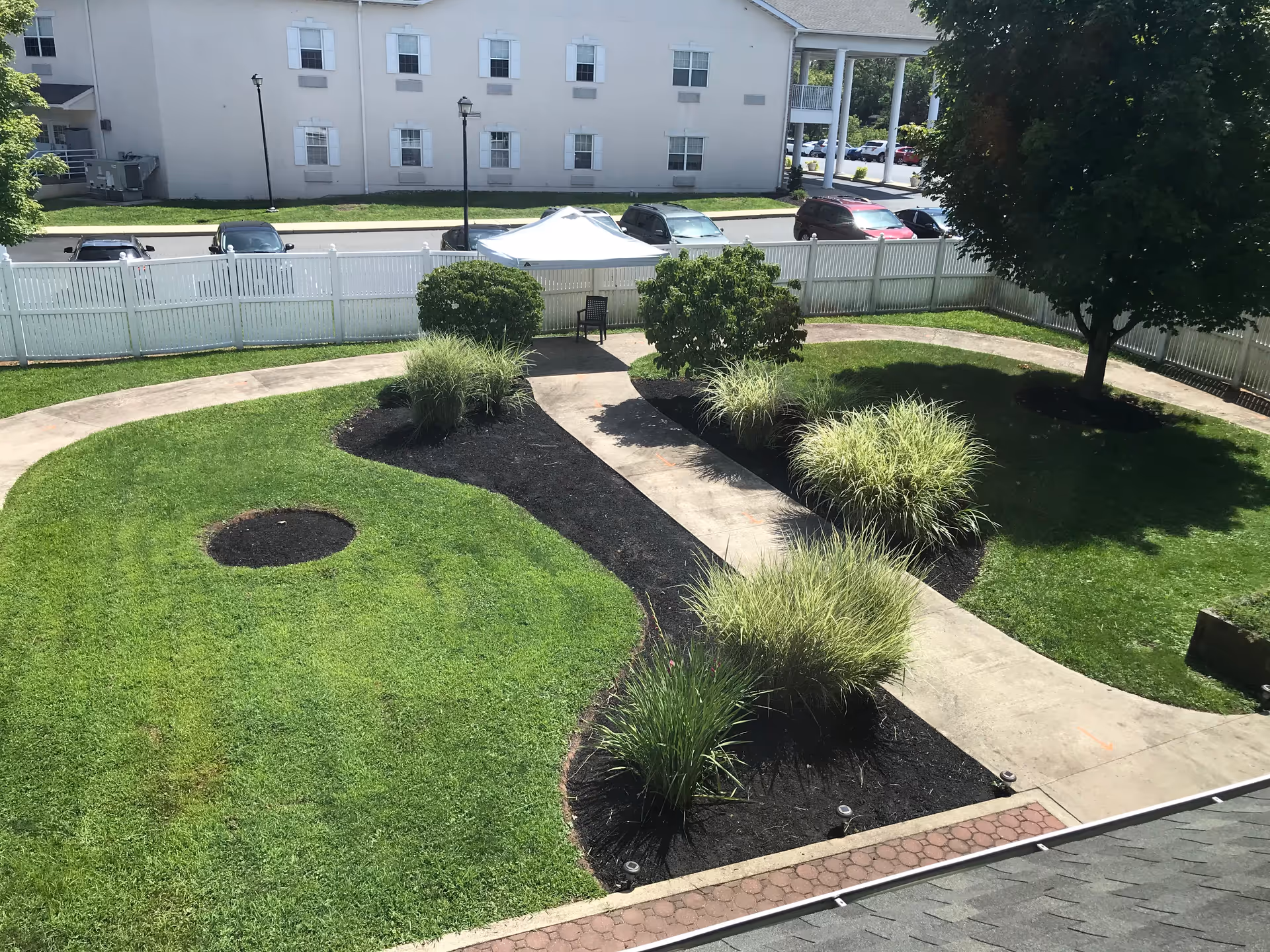 A landscaped outdoor garden area with a curved concrete pathway, green grass, bushes, and a large tree. There is a white fence surrounding the garden, a white canopy tent with a chair underneath, and a building with multiple windows and parked cars visible in the background.