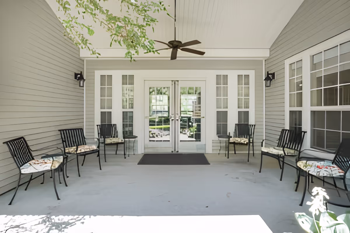 Covered outdoor seating area with six black metal chairs with cushions arranged along the walls. The area has a ceiling fan and is adjacent to a building with large windows and double glass doors.