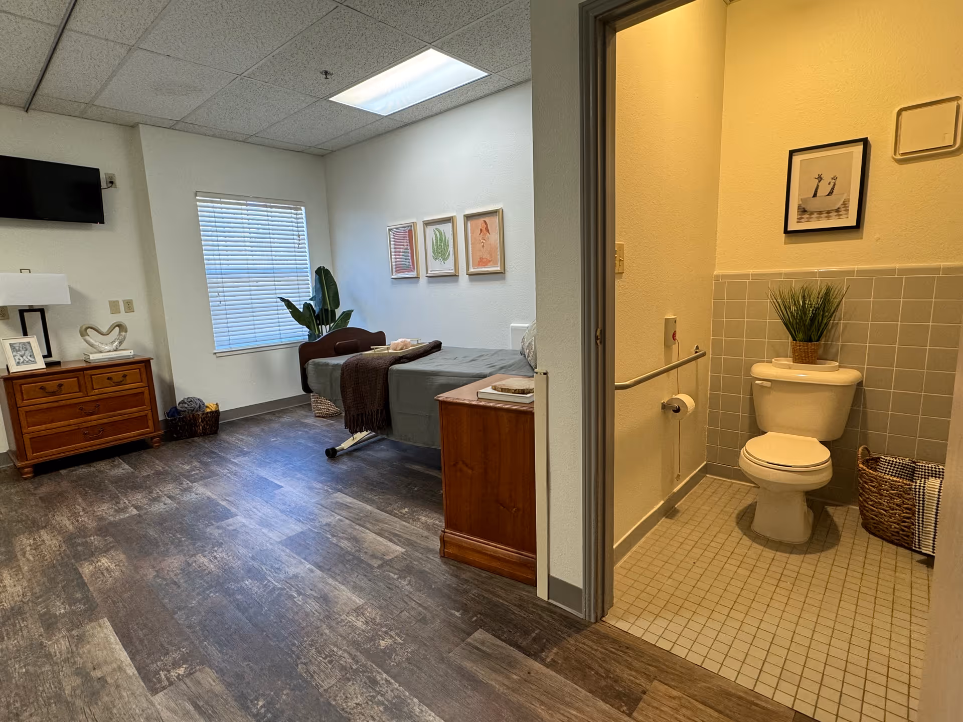 Furnished resident bedroom with a bed, dresser, TV and window, plus an open door showing a tiled bathroom with a toilet and plant.