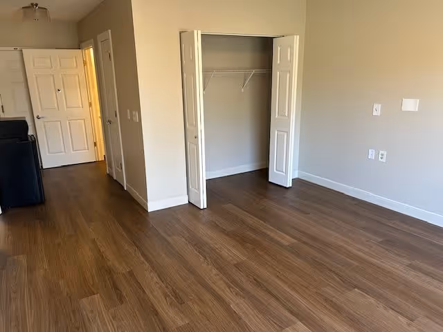 Empty room with wooden flooring, beige walls, an open closet with white bi-fold doors, and a hallway with white doors leading to other rooms.