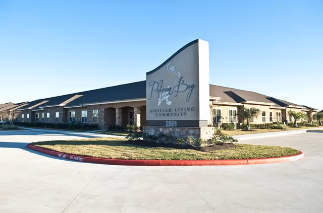 Exterior view of Pelican Bay Assisted Living Community building with a large sign displaying the facility name and address 2501, surrounded by a driveway and landscaped grass area under a clear blue sky.