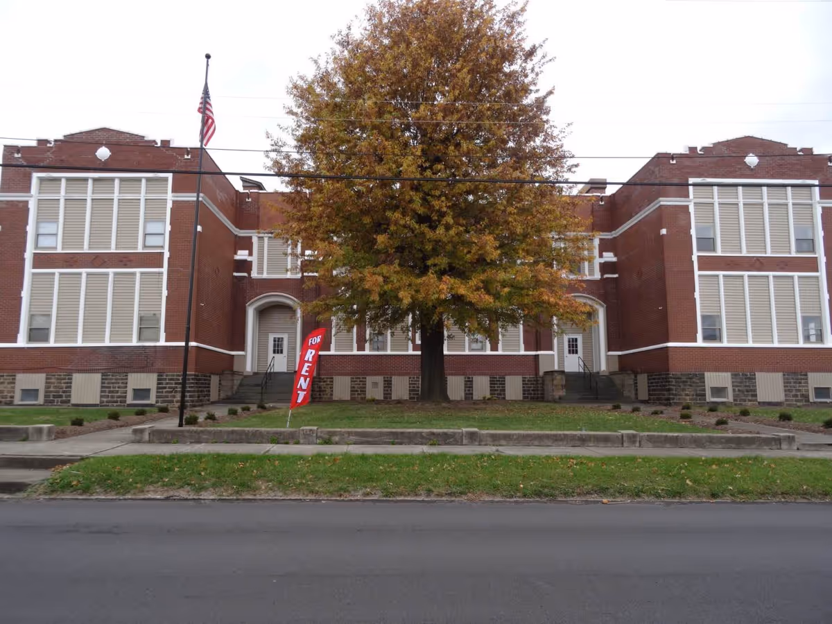 Front exterior view of a large brick building with white window shutters and two main entrances. A large tree with autumn-colored leaves stands in the center of the lawn. An American flag is on a flagpole to the left, and a red 'For Rent' sign is placed near the tree.