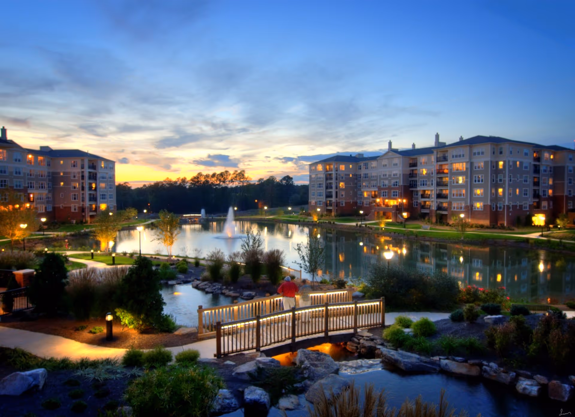 Evening view of a senior living facility with multi-story buildings surrounding a large pond with a fountain. There is a well-lit walking path and a wooden bridge over a small stream in the landscaped garden area. The sky is partly cloudy with the colors of sunset.