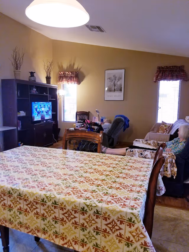 A cozy living area with a dining table covered by a patterned tablecloth in the foreground. In the background, there is a TV on a dark wooden entertainment center, two windows with plaid valances, a recliner chair, and a sofa where an elderly person is seated. The walls are painted beige and decorated with a framed picture.