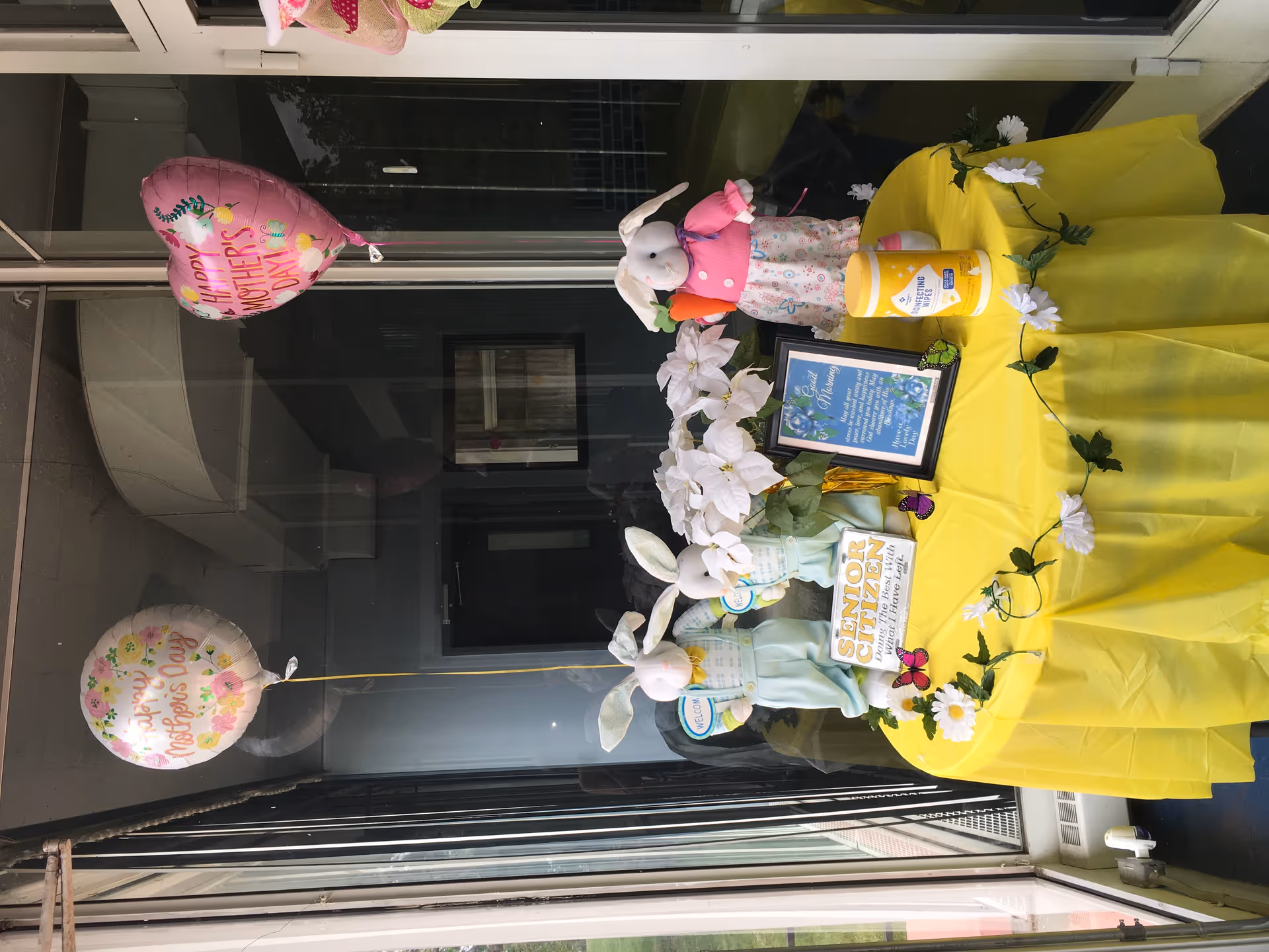 A small round table covered with a yellow tablecloth is decorated with two stuffed bunny dolls, white artificial flowers, butterfly decorations, and two balloons that say 'Happy Mother's Day'. One bunny is holding a carrot and the other two have 'Welcome' signs. There is a framed message on the table and a container of disinfecting wipes. The table is placed near a glass window and door.