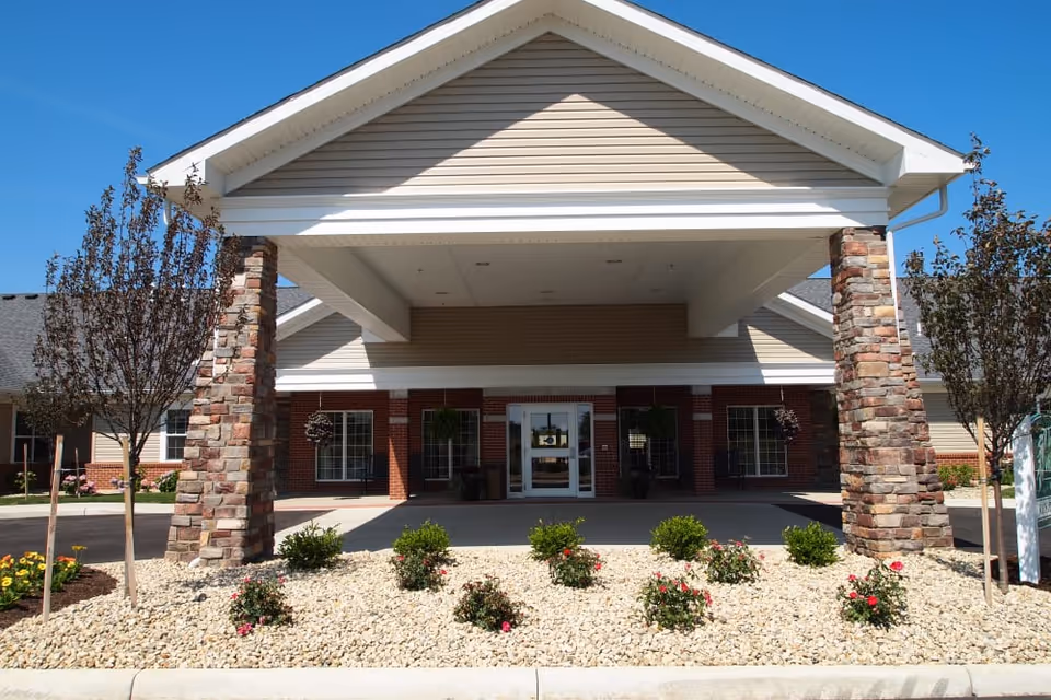 Covered entrance of a senior living facility with stone pillars, rock landscaping and glass double doors.
