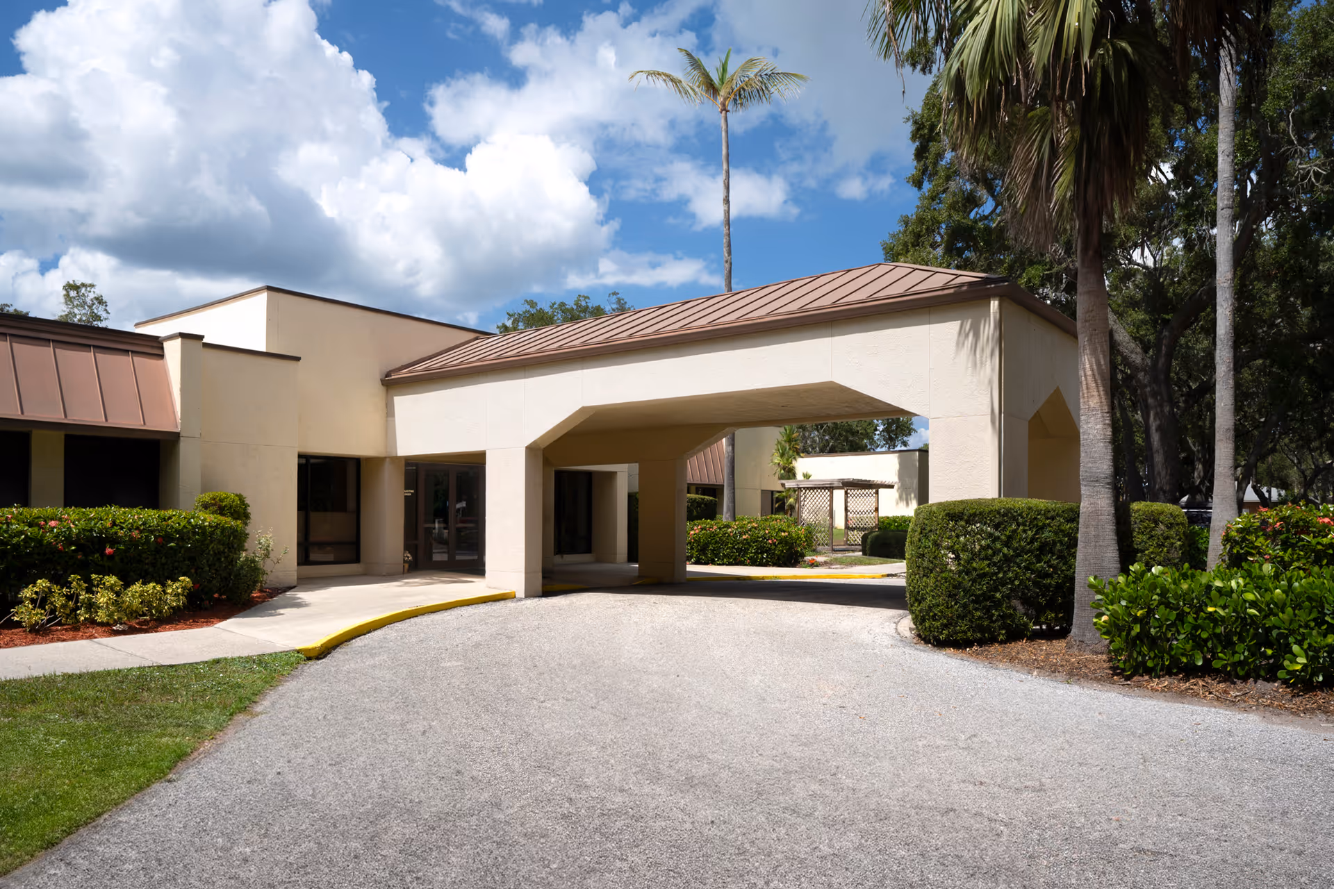 Exterior view of Heritage Park Health Center showing a beige building with a covered entrance driveway, surrounded by trimmed bushes, palm trees, and a partly cloudy sky.