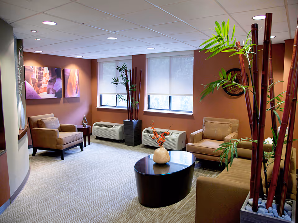 A cozy living room area with beige armchairs and a sofa arranged around a dark oval coffee table with a decorative vase. The walls are painted in warm tones, and there are two windows with white blinds. Tall plants in modern planters add greenery to the space, and framed artwork hangs on the wall.