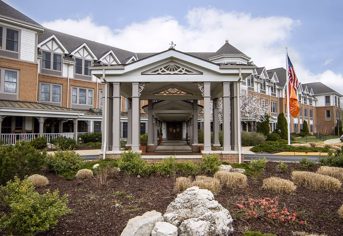 Front exterior view of Sunrise of Des Peres senior living facility featuring a covered entrance with columns, landscaped garden with shrubs and rocks, and flags including an American flag and an orange flag.