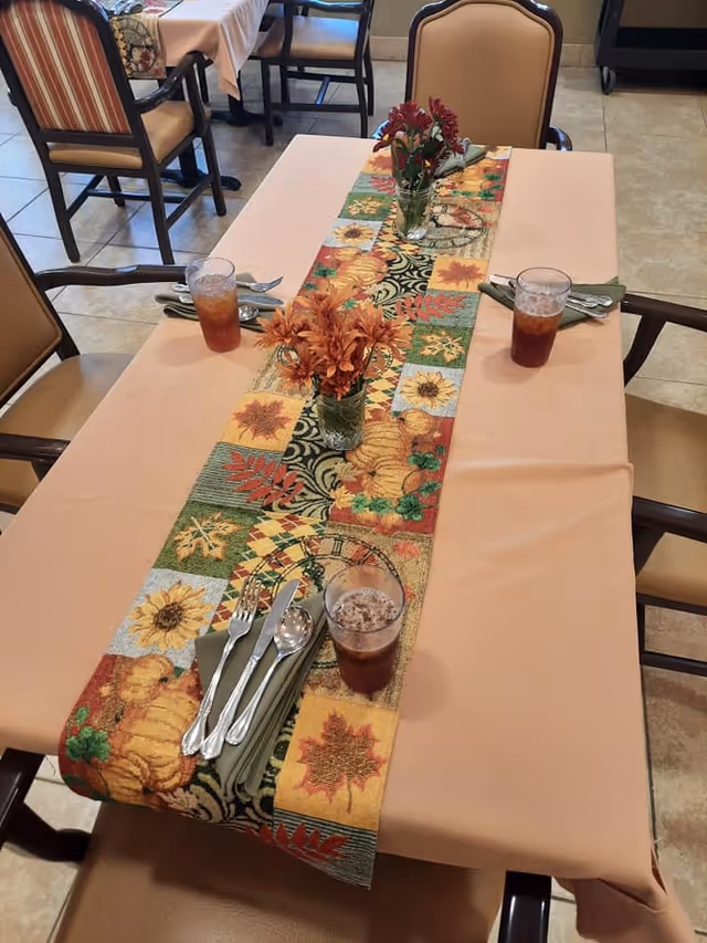 A dining table set with a beige tablecloth and a colorful autumn-themed table runner featuring sunflowers, leaves, and pumpkins. There are three glasses of iced tea, three sets of silverware wrapped in green napkins, and two small vases with orange flowers on the table. Surrounding the table are cushioned chairs with wooden frames. The floor is tiled.