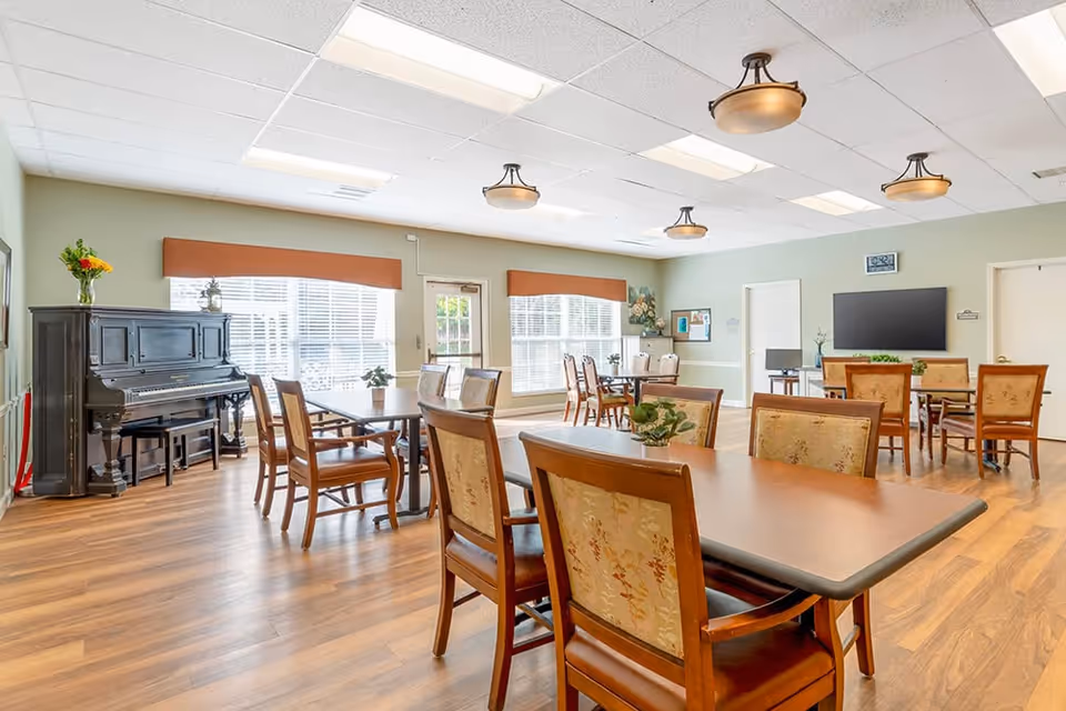 Bright communal dining/activity room with multiple tables and chairs, a piano, TV, and large windows.