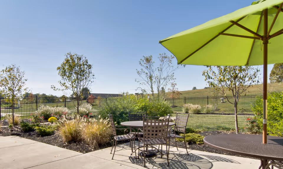 Patio with metal tables and chairs under a green umbrella overlooking landscaped gardens, lawn and a fenced open field.