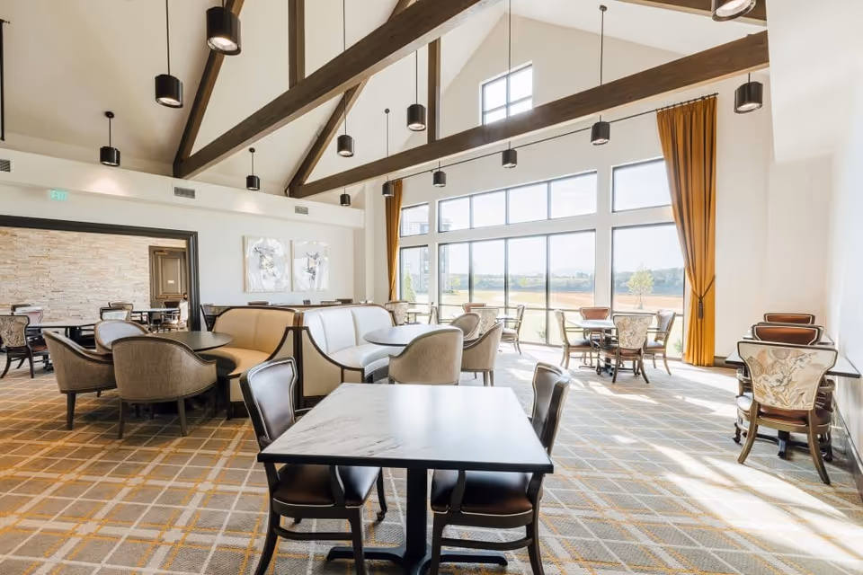Bright and spacious dining area with large windows letting in natural light, featuring multiple tables and chairs arranged for seating. The room has high vaulted ceilings with exposed wooden beams and modern pendant lights. There are mustard yellow curtains on the windows and a patterned carpet on the floor.