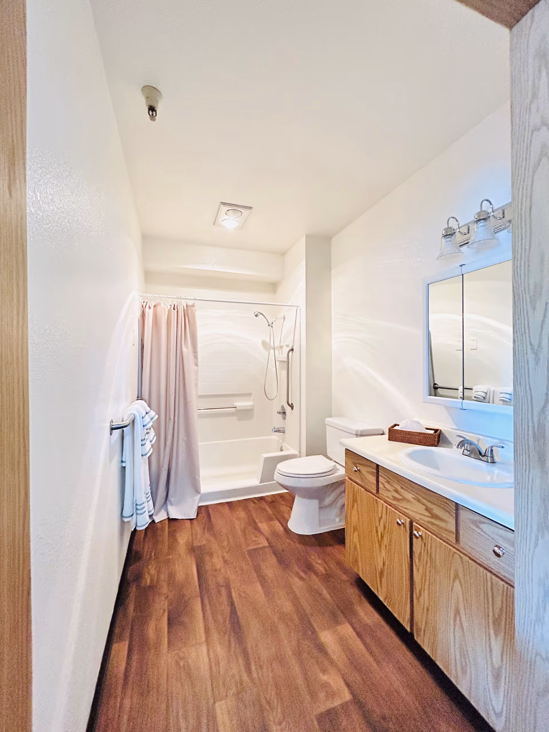 A clean bathroom with wood-look flooring, a white bathtub with a shower and pink curtain, a white toilet, a wooden vanity with a white sink, a mirror with lights above it, and towels hanging on a rack on the left wall.