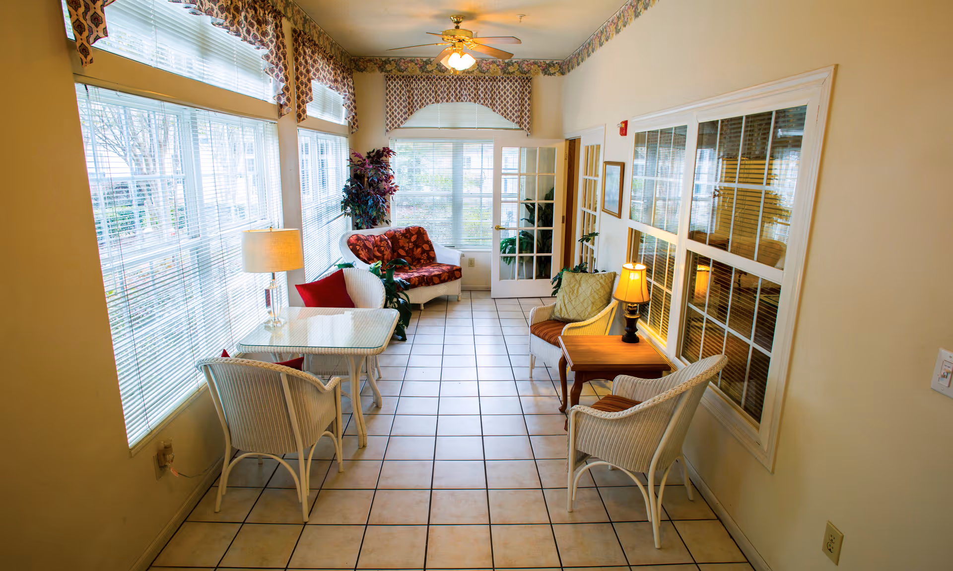 A bright sunroom with large windows on the left side covered with blinds and patterned valances. The room features white wicker furniture including chairs and a loveseat with red and orange cushions, two small tables with lamps, and several potted plants. The floor is tiled, and there is a ceiling fan with lights. A glass door and a large window are on the far end and right side of the room.