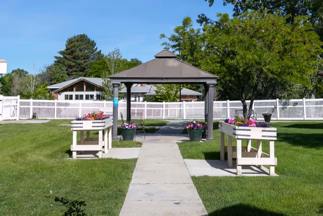 Outdoor garden area with a concrete pathway leading to a gazebo structure. There are raised flower beds on either side of the pathway filled with colorful flowers. The area is surrounded by green grass, trees, and a white fence with buildings visible in the background under a clear blue sky.