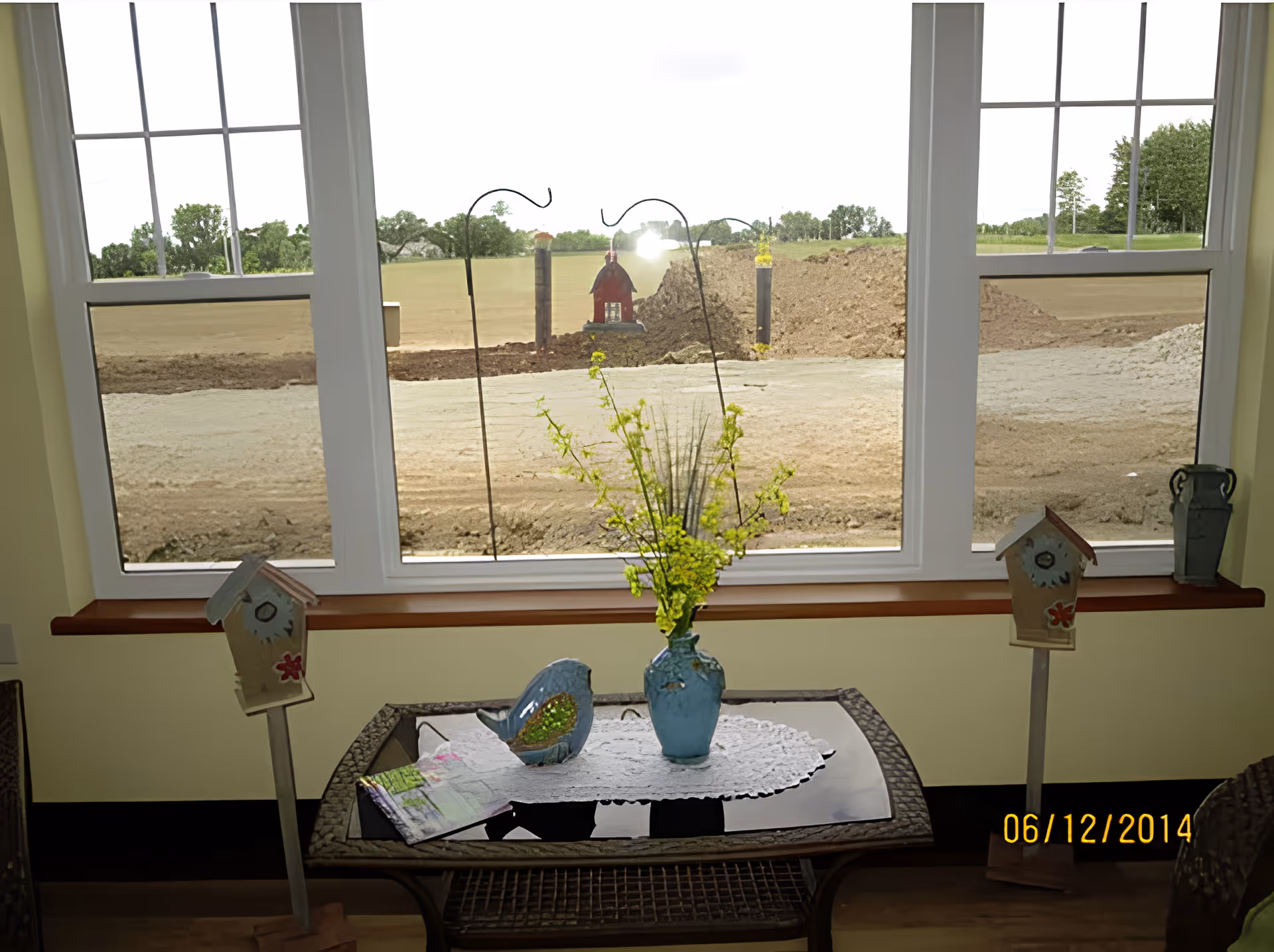 A small indoor table with a glass top and wicker frame is placed in front of a large window. On the table, there is a blue vase with yellow flowers, a decorative blue bird, and a piece of paper on a white doily. Outside the window, there is a dirt area with some construction or landscaping work in progress, and green trees in the distance.