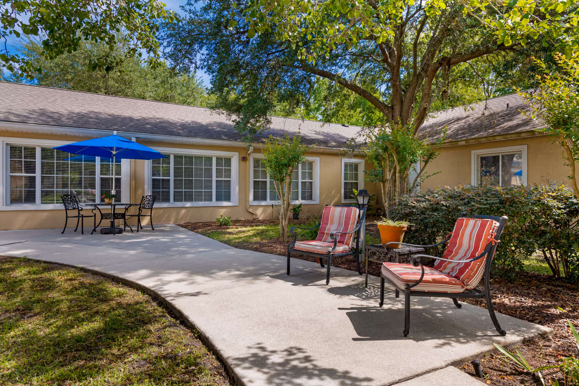 Outdoor patio area at a senior living facility with two cushioned chairs and a small table under a large tree. In the background, there is a building with multiple windows and a table with four chairs shaded by a blue umbrella. The area is surrounded by greenery and bushes under a clear blue sky.