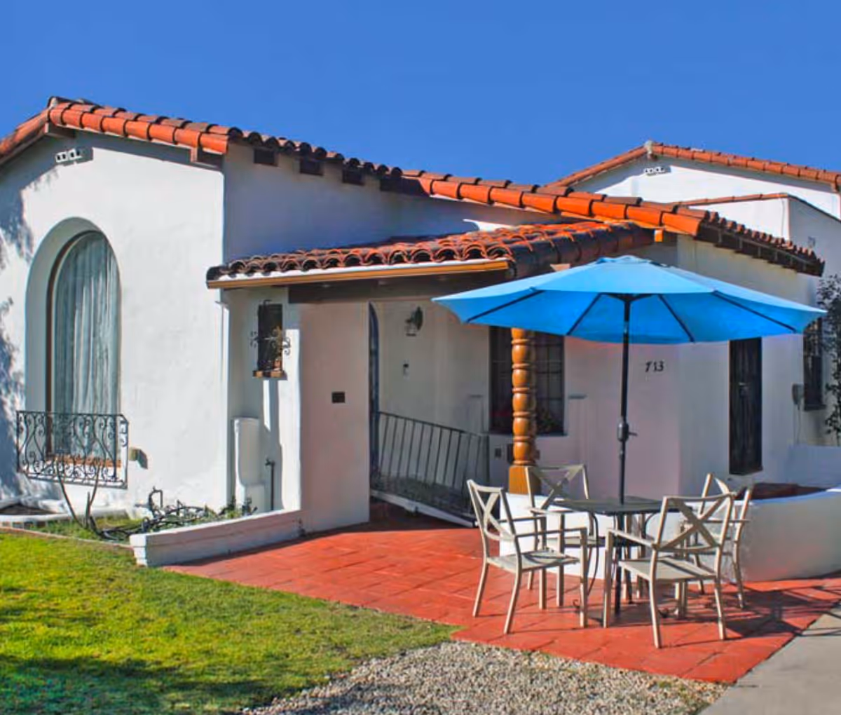 Exterior view of a white stucco building with a red tile roof under a clear blue sky. In front of the building, there is a patio area with a glass-top table, four chairs, and a large blue umbrella providing shade. The building has an arched window with decorative ironwork and a small ramp leading to the entrance.