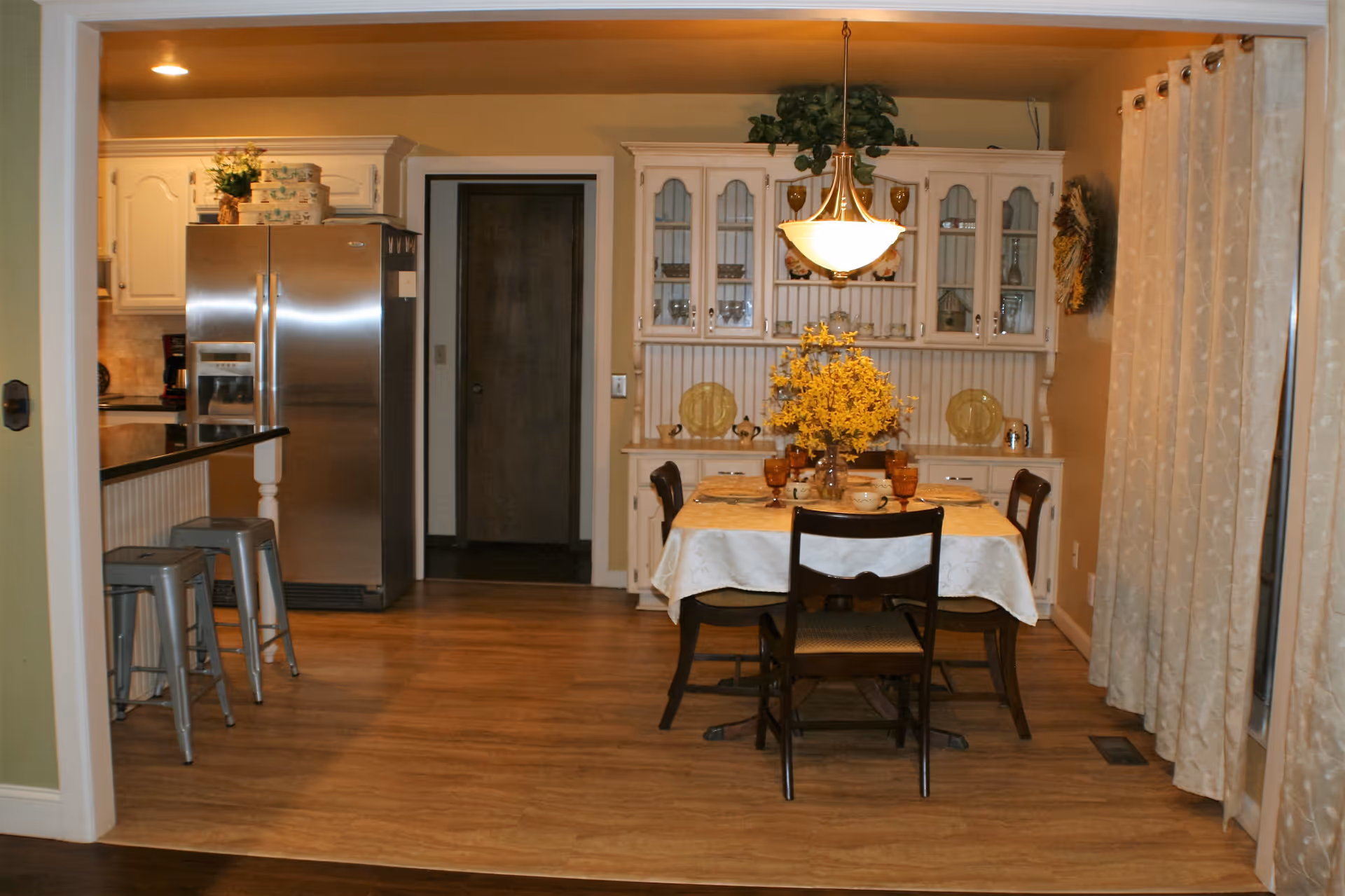 A cozy dining area with a table set for four, featuring a white tablecloth and a centerpiece of yellow flowers. Behind the table is a white hutch with glass doors displaying dishes and decorative items. To the left, a kitchen area is visible with a stainless steel refrigerator and two metal bar stools at a counter. Beige curtains cover the window on the right side of the room.