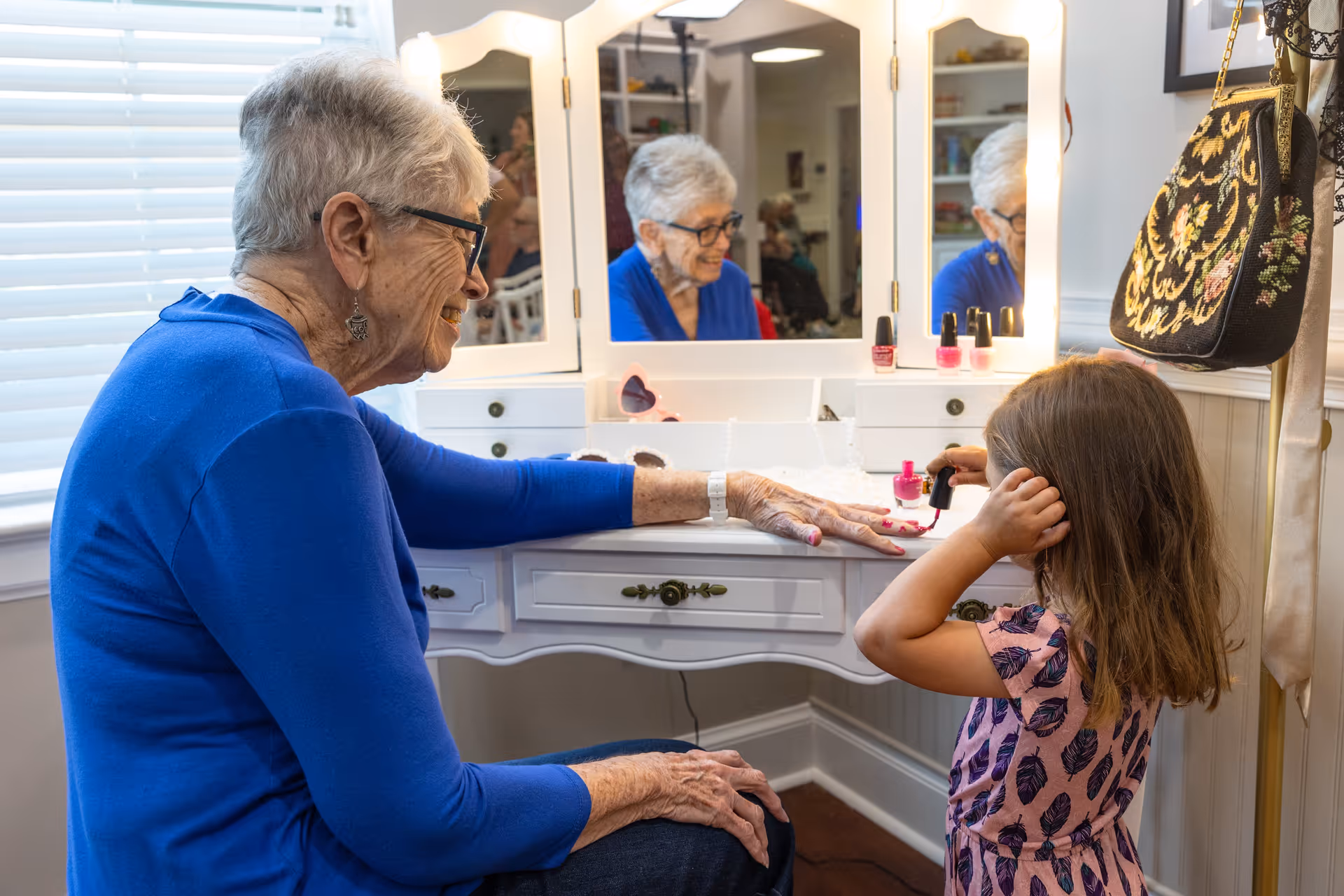An elderly woman wearing glasses and a blue sweater is sitting at a white vanity table while a young girl in a pink dress with feather patterns paints the woman's fingernails with pink nail polish. The vanity has a mirror with lights and several bottles of nail polish on it. A decorative handbag hangs on the wall beside the vanity.