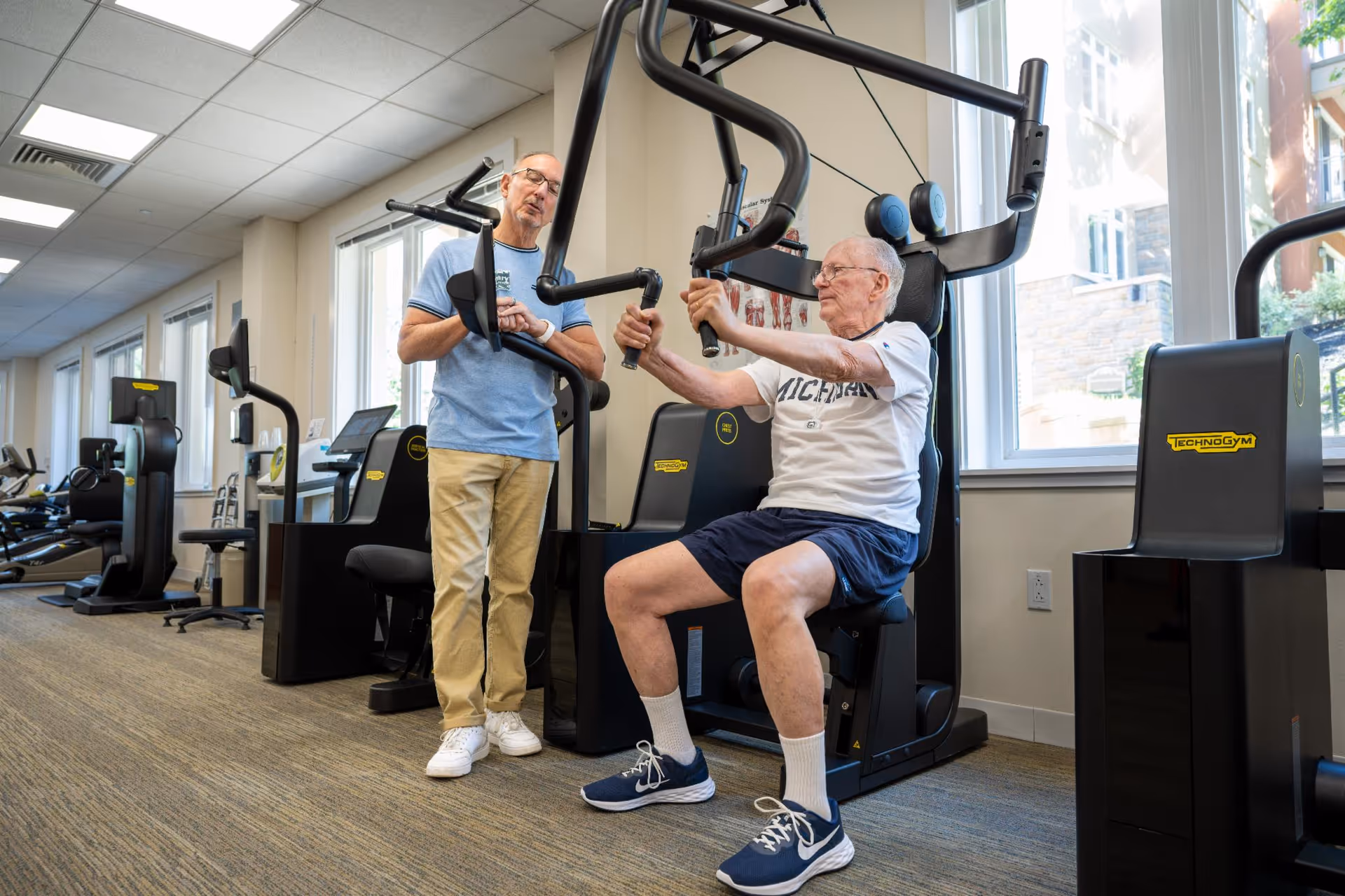 An elderly man wearing a Michigan t-shirt and shorts is using a chest press exercise machine in a gym. Another man in a blue shirt and beige pants stands nearby, watching and smiling. The gym has several exercise machines, large windows letting in natural light, and a carpeted floor.